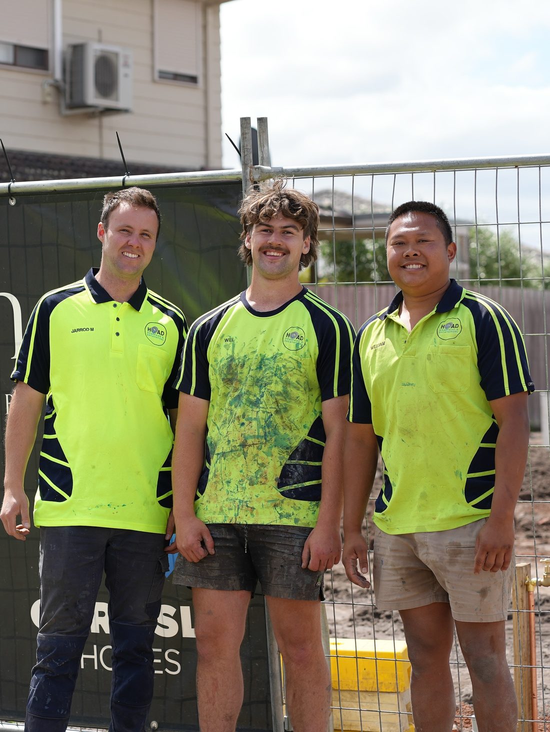 Three Hoad Drainage and Excavations team members in branded hi-vis polos in front of site fencing.