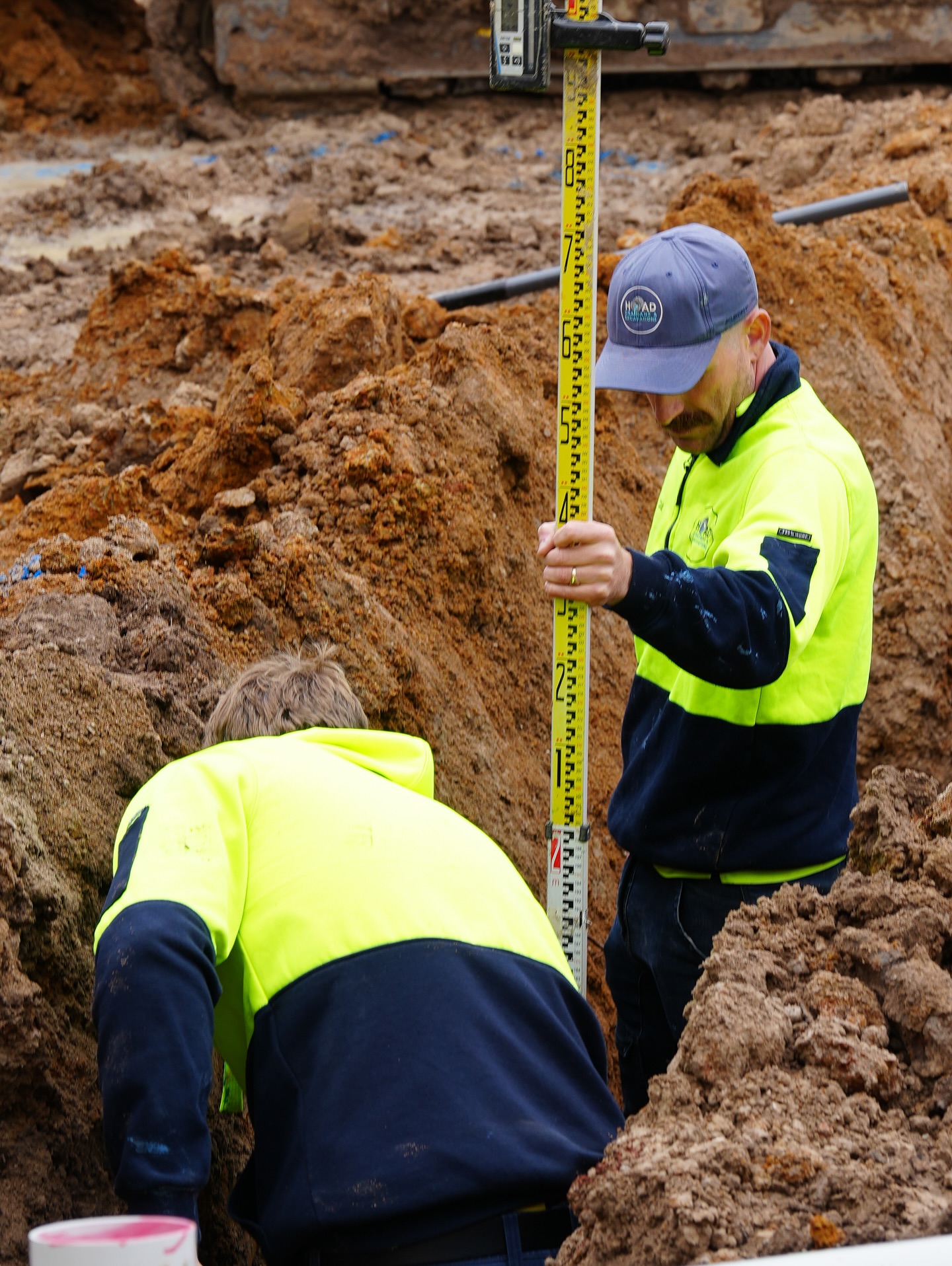 Two crew members in hi-vis taking a level read with a survey staff during a trench dig.